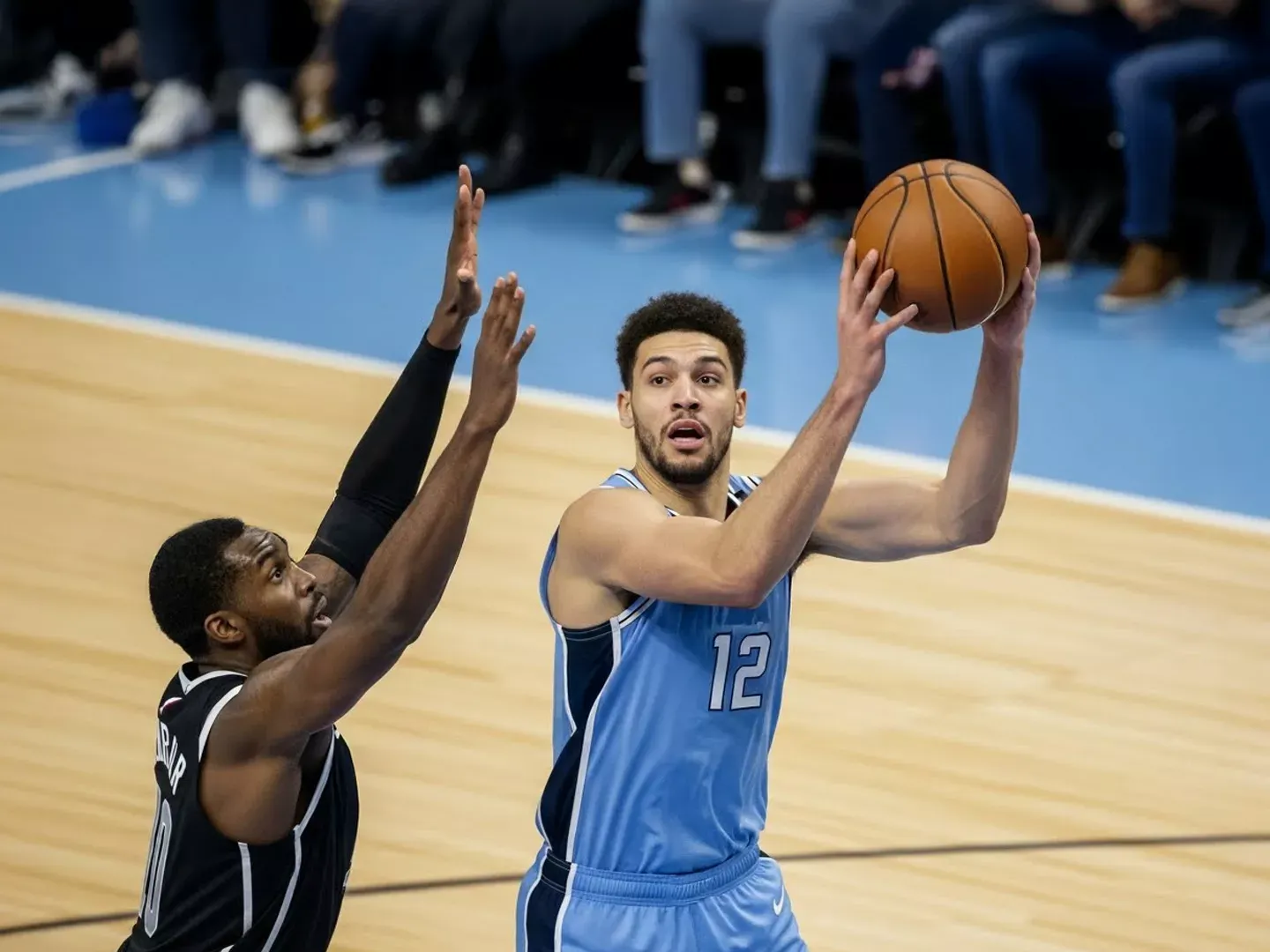 A basketball player in a light blue jersey with the number 12 holds a basketball, preparing to shoot over a defending player with their hands raised.