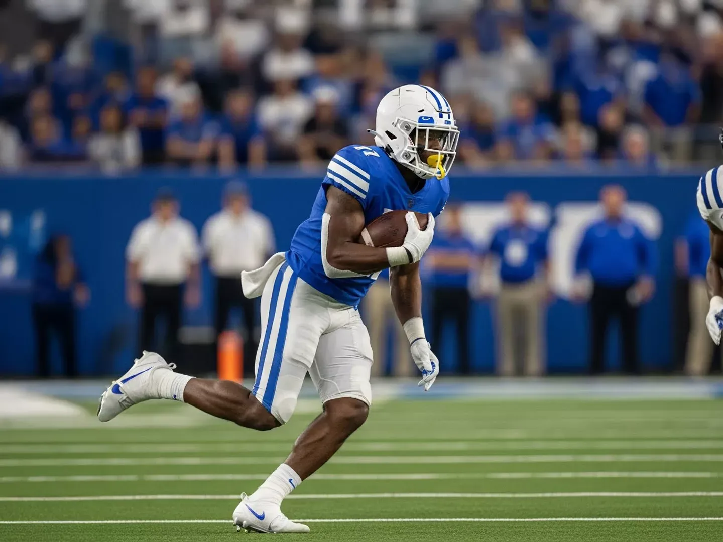 A college football player in a blue and white jersey runs across the field while carrying the football.