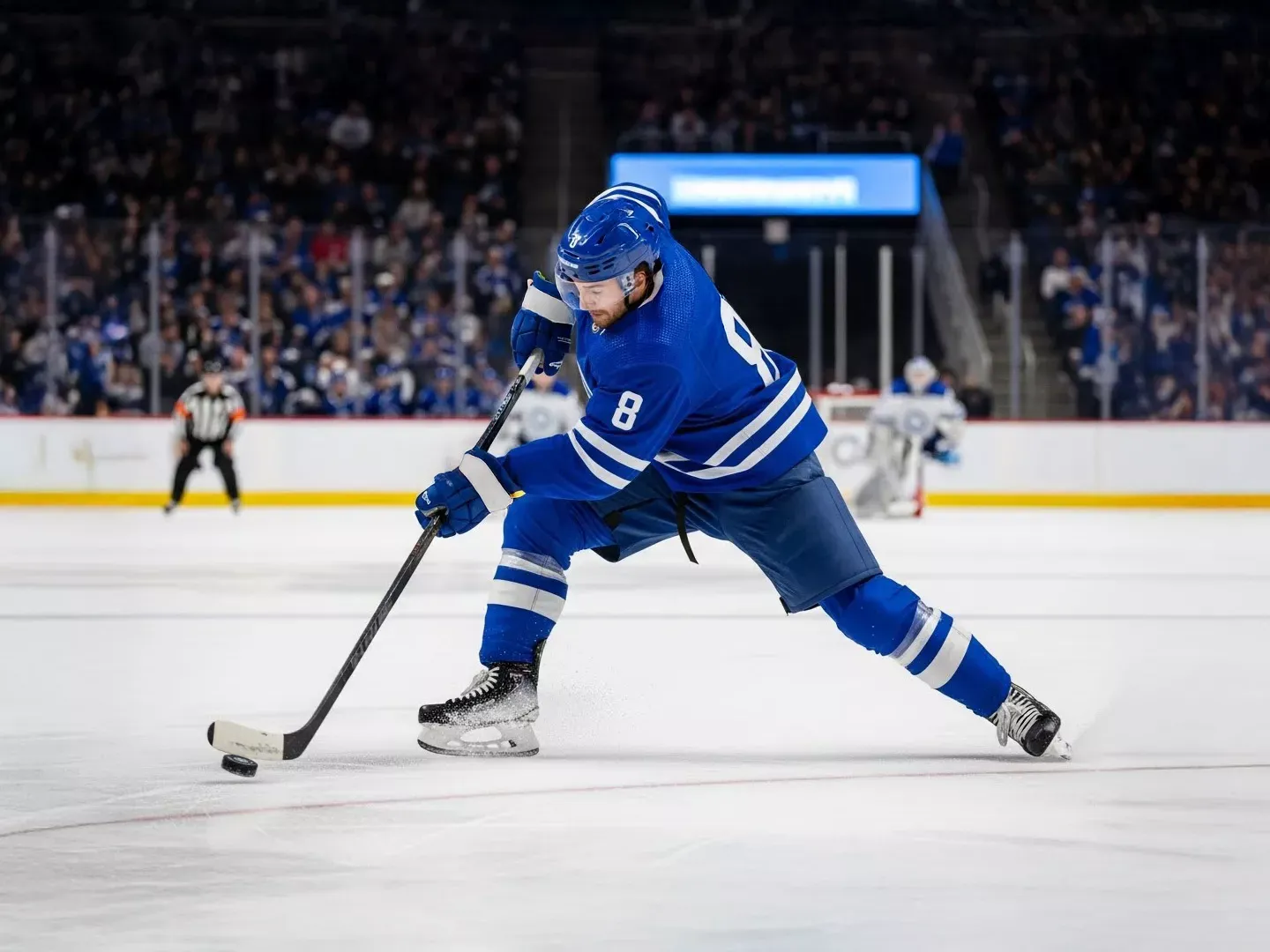 An ice hockey player in a blue jersey with the number 8 takes a slapshot with the puck on the ice in a crowded arena.