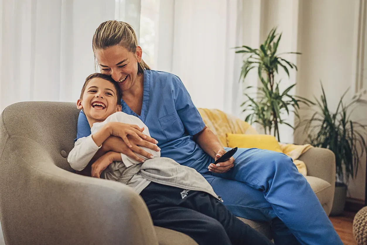 A woman in blue scrubs hugs a laughing boy on an armchair, holding a remote.
