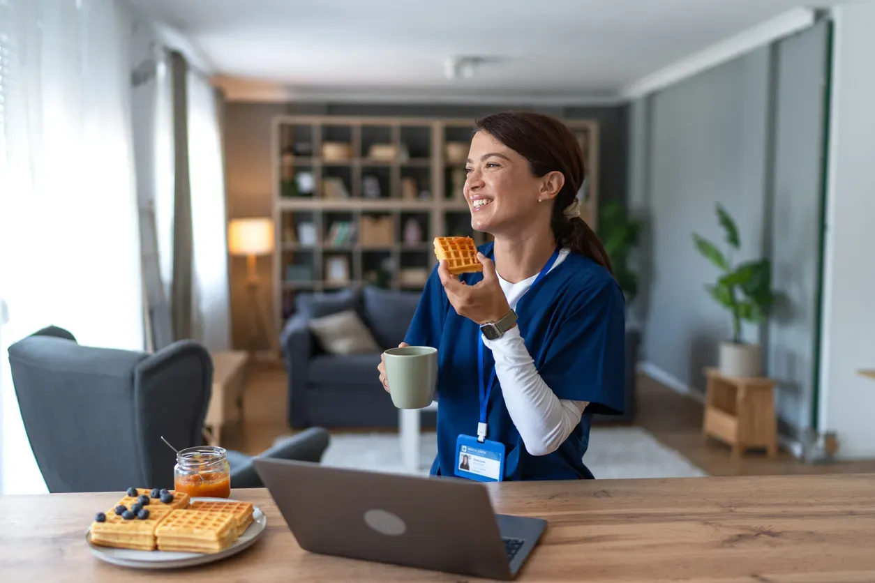 A smiling woman in navy scrubs eats a waffle and holds a mug at home.