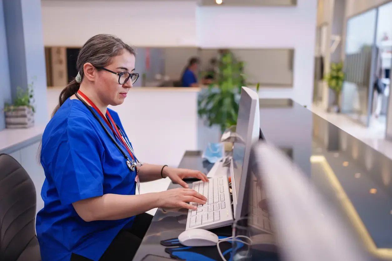 A nurse in blue scrubs types on a keyboard at a clinic reception desk.