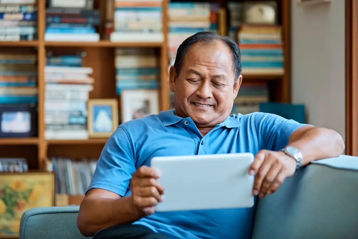 An older man in a blue polo smiles while reading a tablet beside a bookshelf.