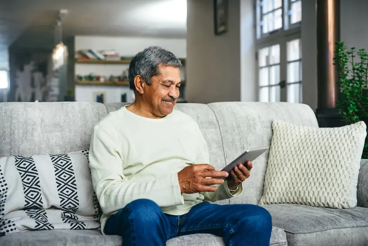 A smiling older man uses a tablet while relaxing on a grey couch at home.
