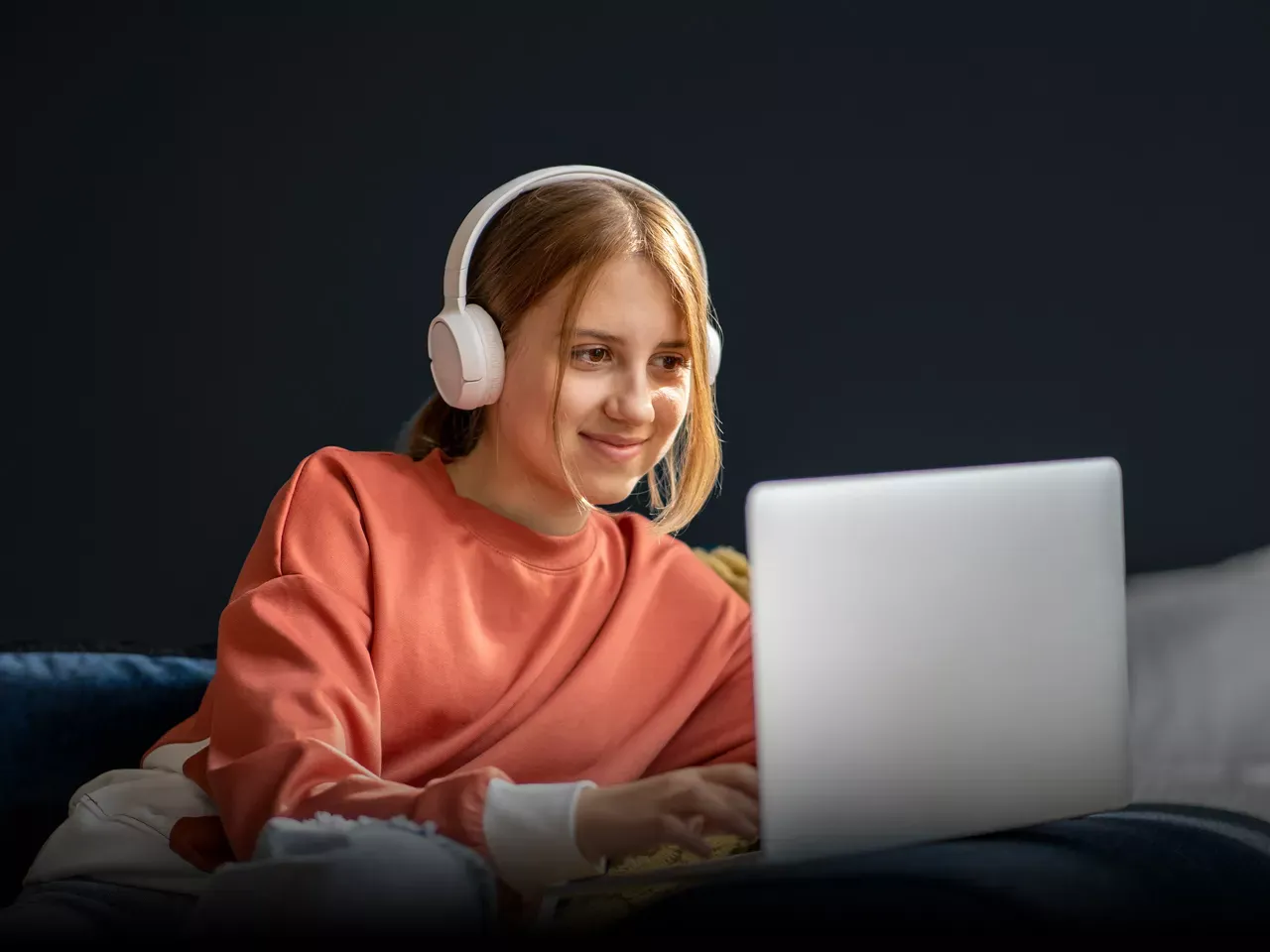 A young woman wearing white headphones and a salmon-colored sweater uses a laptop in a dimly lit room.
