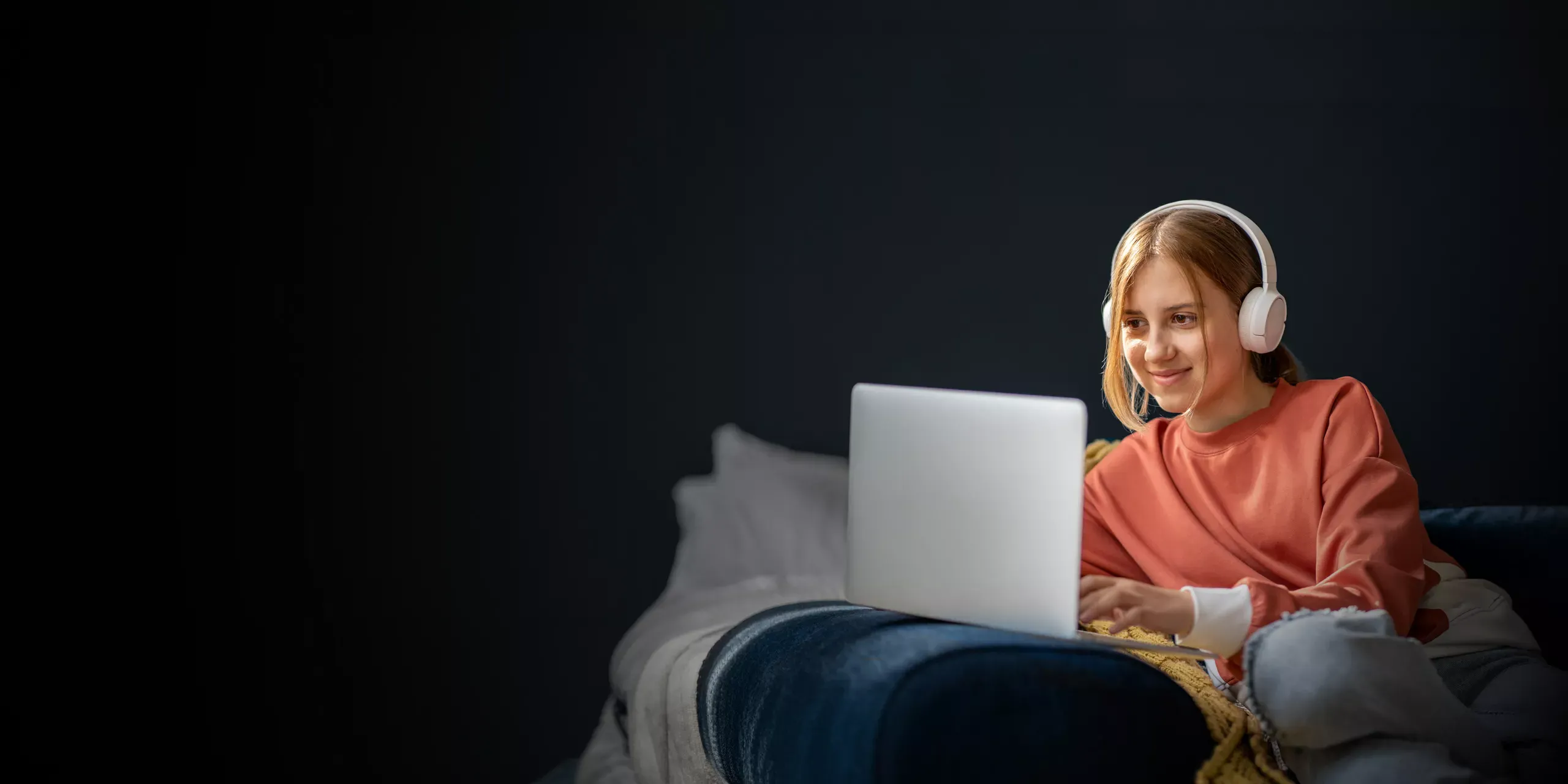 A young woman wearing white headphones and a salmon-colored sweater uses a laptop in a dimly lit room.
