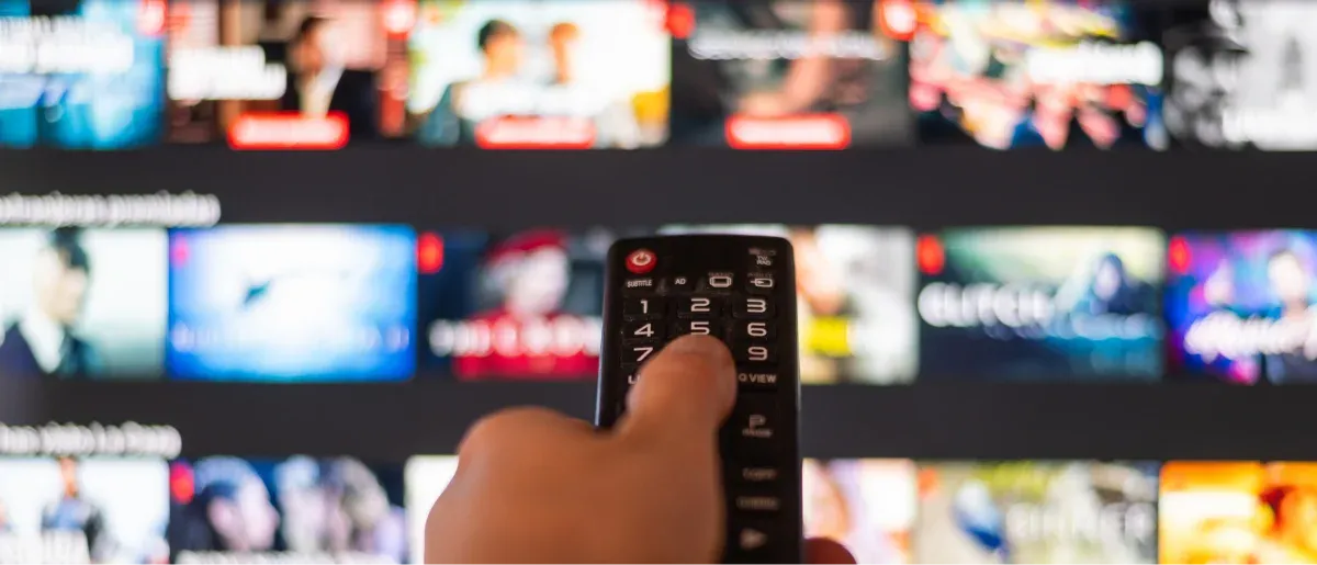 A hand points a remote at a TV screen full of streaming show thumbnails.