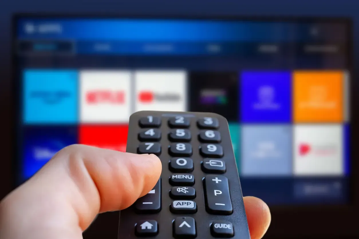 A hand points a black remote at a smart TV displaying a colorful streaming app menu.