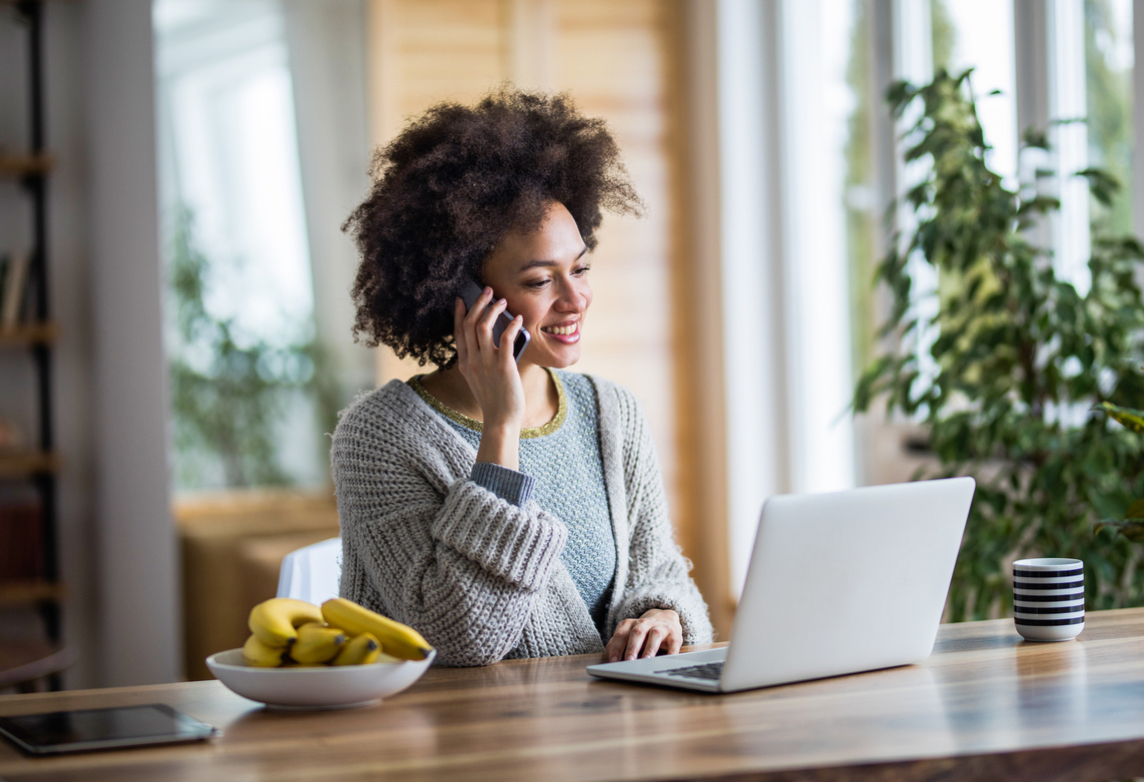 woman talks on the phone and works on a laptop at a table.