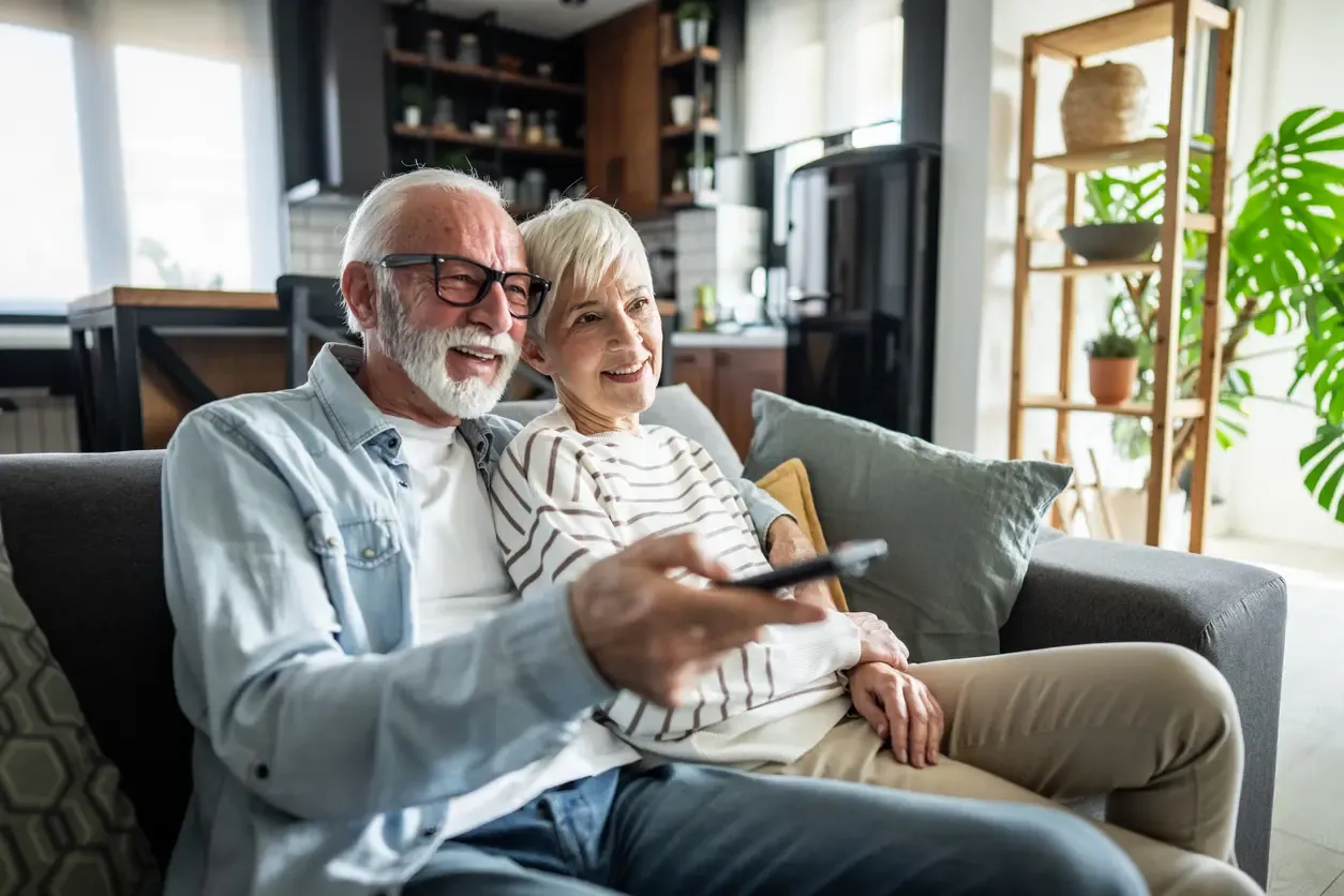 A smiling senior couple sits close on a gray couch while pointing a remote at the television.