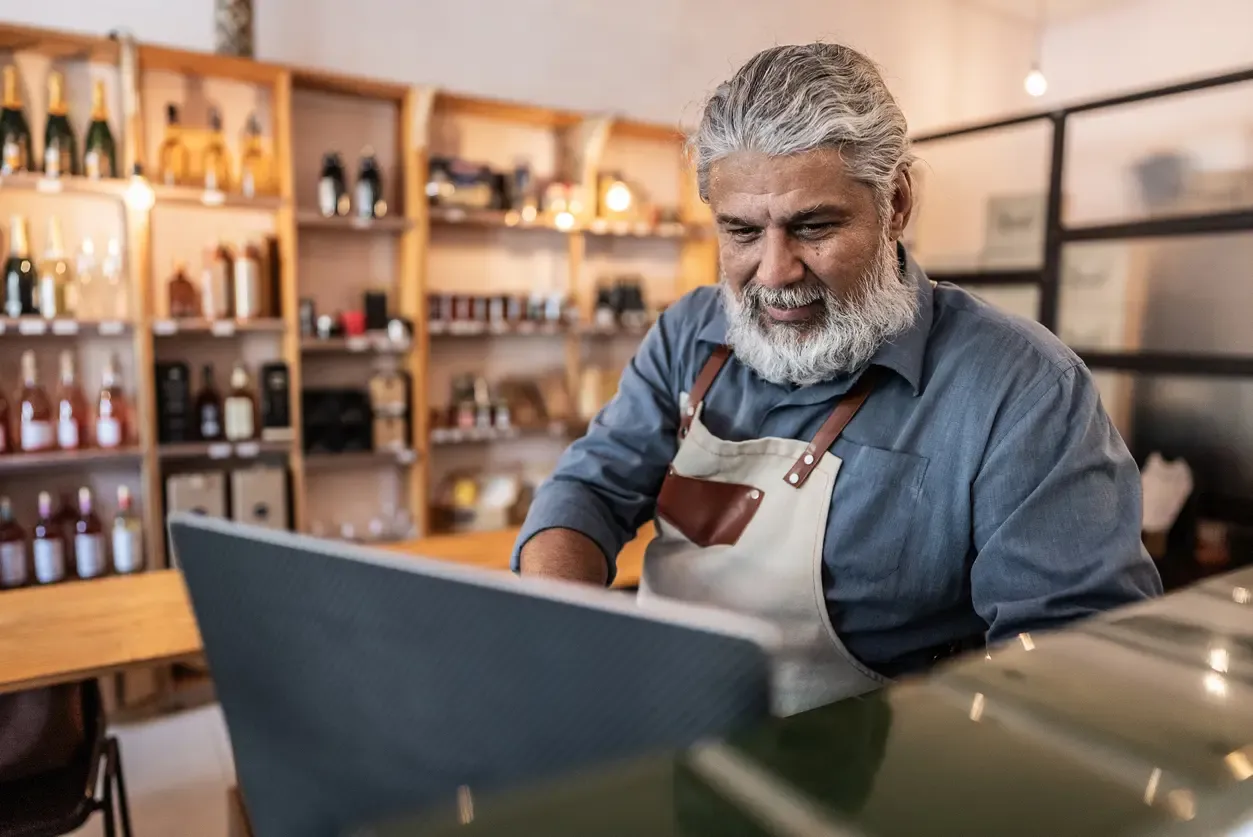 A senior male shop owner with a gray beard and apron looks down at a laptop screen in a boutique store with wine shelves in the background.