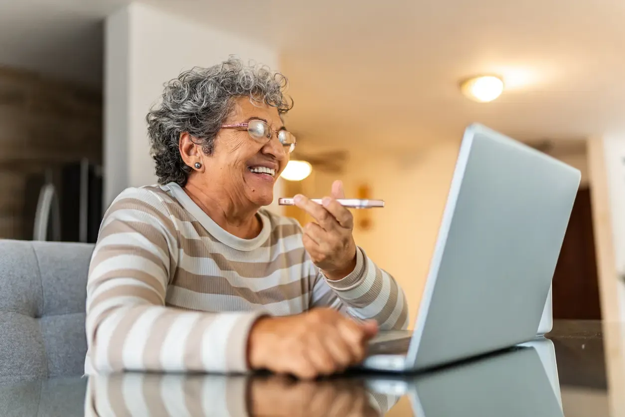 A smiling older woman with glasses holds a phone while using a laptop at a glass table at home.