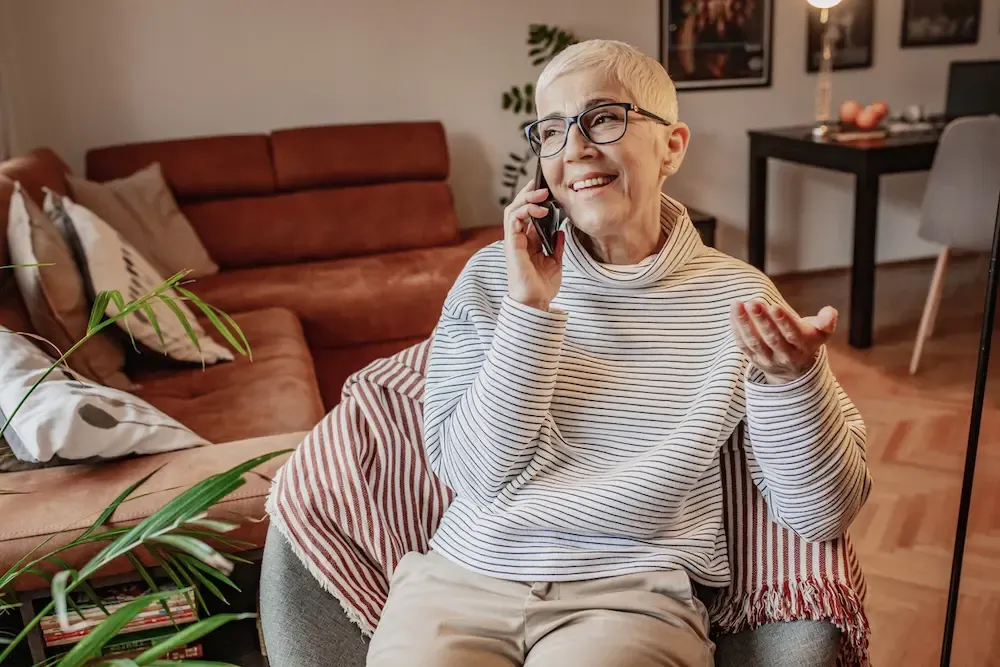 A smiling older woman in a striped sweater chats on her phone in a cozy living room