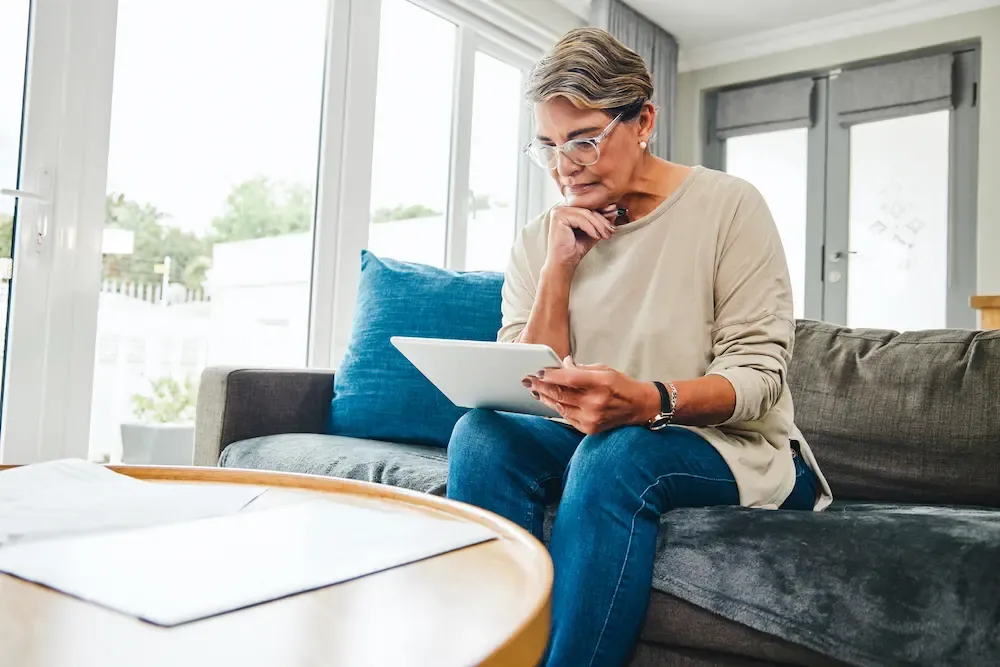 A senior woman with glasses sitting on a sofa and looking thoughtfully at a digital tablet.