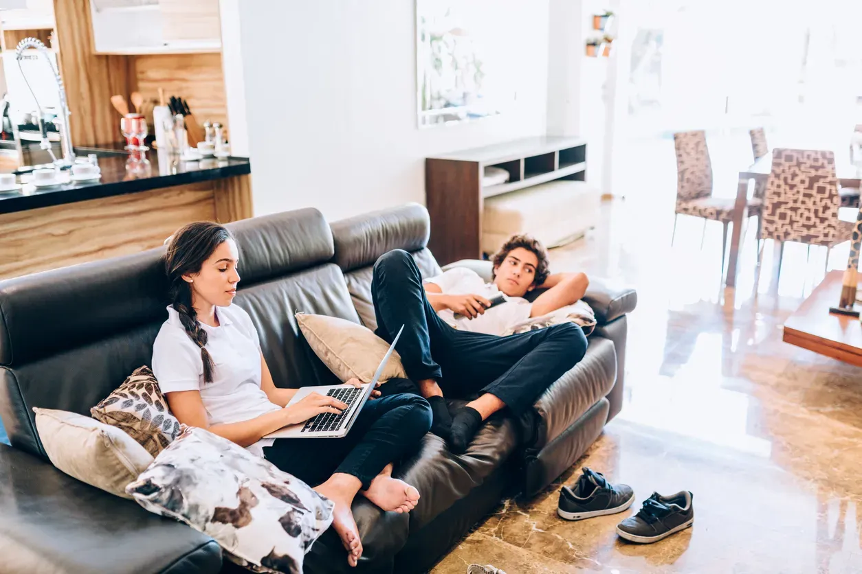 A young woman types on a laptop while a man reclines nearby holding a remote.