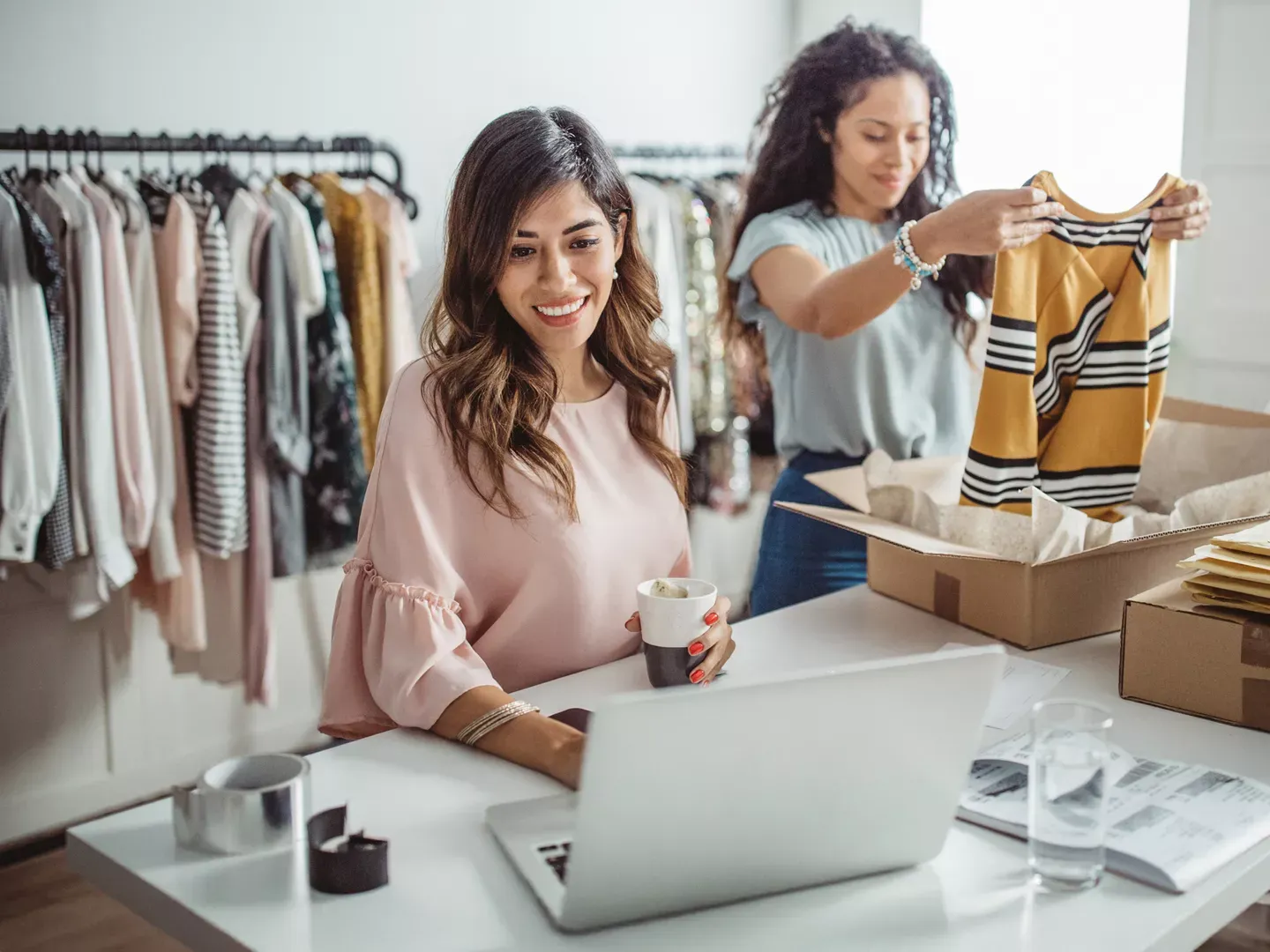 A woman uses a laptop and holds a drink while another woman packs clothing into a box at a small business.