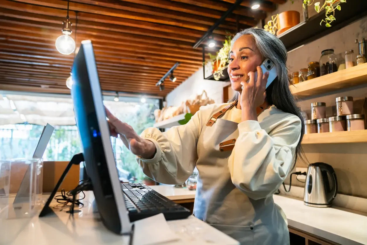 A smiling woman with gray hair talks on a smartphone while using a computer at a shop counter.