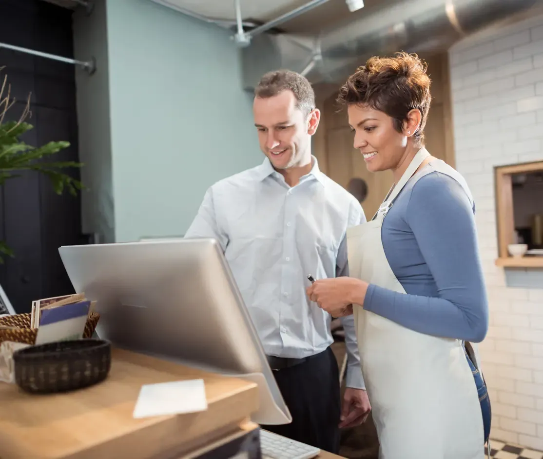 Two small business owners smile while reviewing information on a computer screen at a counter.