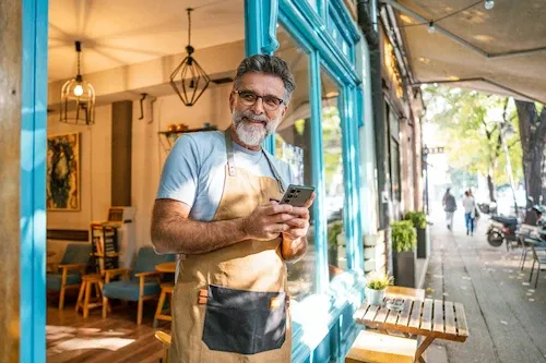 Smiling mature man wearing glasses and an apron standing in the doorway of his small business while holding a smartphone.