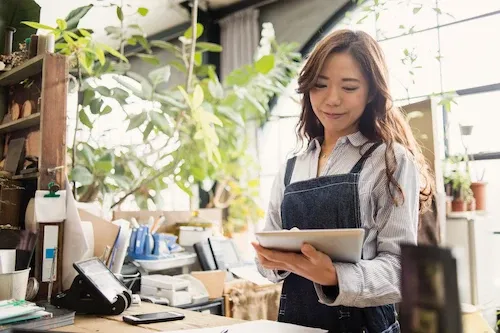 Young woman wearing an apron and striped shirt working in a bright, plant-filled shop while looking at a digital tablet.
