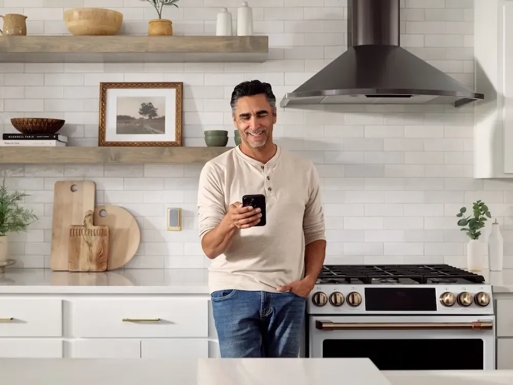 A smiling man in a modern kitchen looks at his smartphone with a chime extender plugged into a wall outlet in the background.