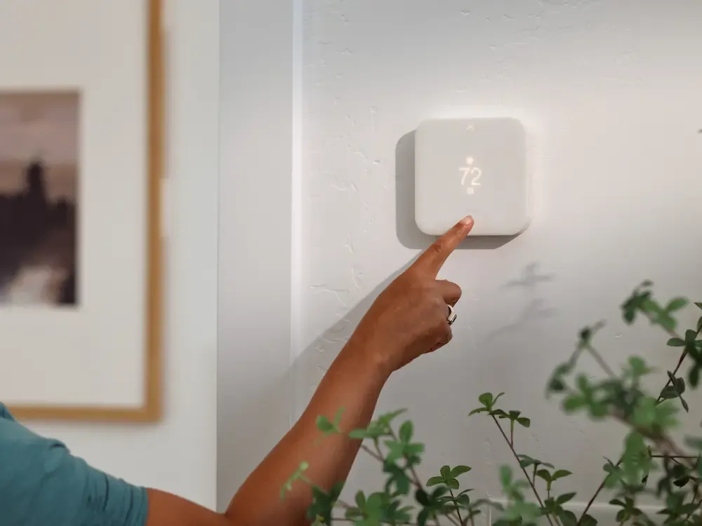 A person's hand touching a white square smart thermostat displaying seventy-two degrees on a white wall.