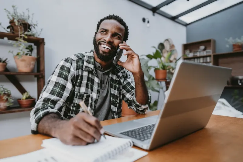A smiling man talks on a cell phone and takes notes in a notebook while sitting at a desk with an open laptop in a plant-filled room.