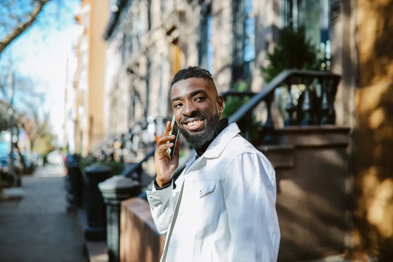 A smiling man with a beard talks on a smartphone while standing on a sunny city sidewalk in front of brownstone buildings.