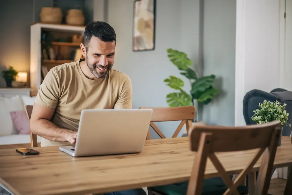 A smiling man sits at a wooden dining table and works on a laptop in a modern living space.
