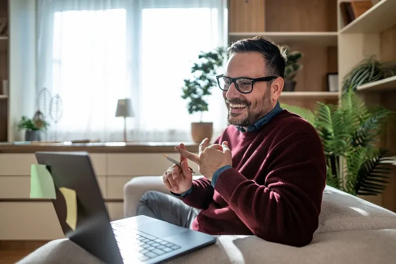A smiling man with glasses sits on a sofa, gesturing with his hand while looking at a laptop screen with sticky notes attached.