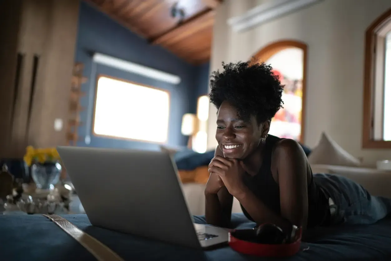 A smiling young woman with an afro hairstyle lies on her stomach on a bed while looking at a laptop screen.