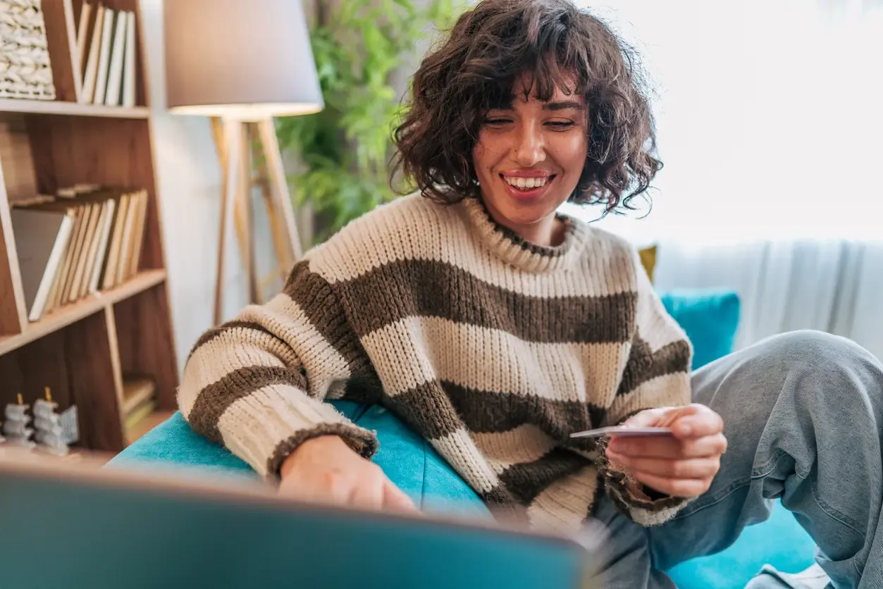 A smiling woman with curly hair wearing a striped sweater sitting on a teal sofa while holding a credit card and using a laptop for online shopping.