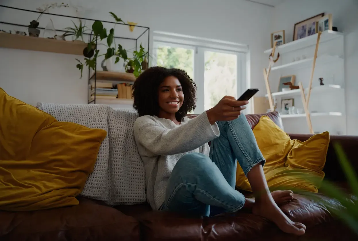 A smiling woman sits barefoot on a brown leather sofa pointing a TV remote in a bright living room.