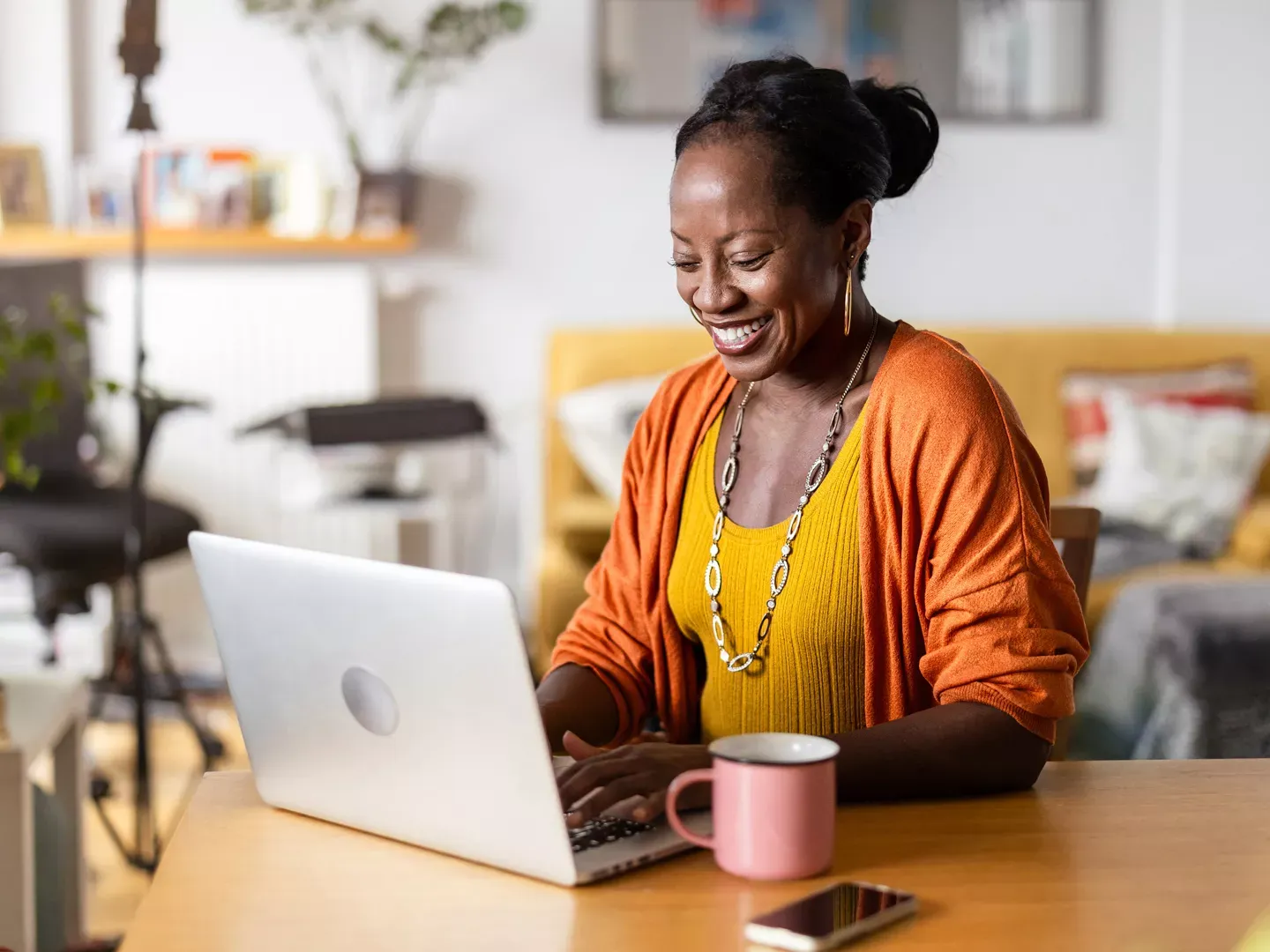 A smiling woman wearing an orange cardigan typing on a laptop at a wooden desk with a coffee mug nearby.