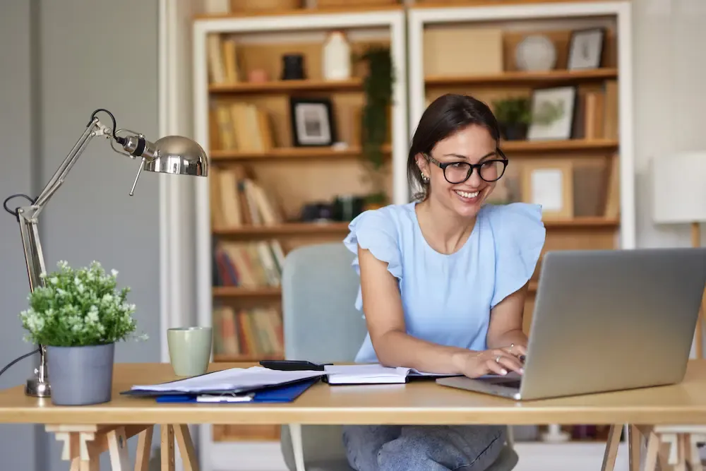 A smiling woman with glasses works on a laptop at a wooden desk in a home office.