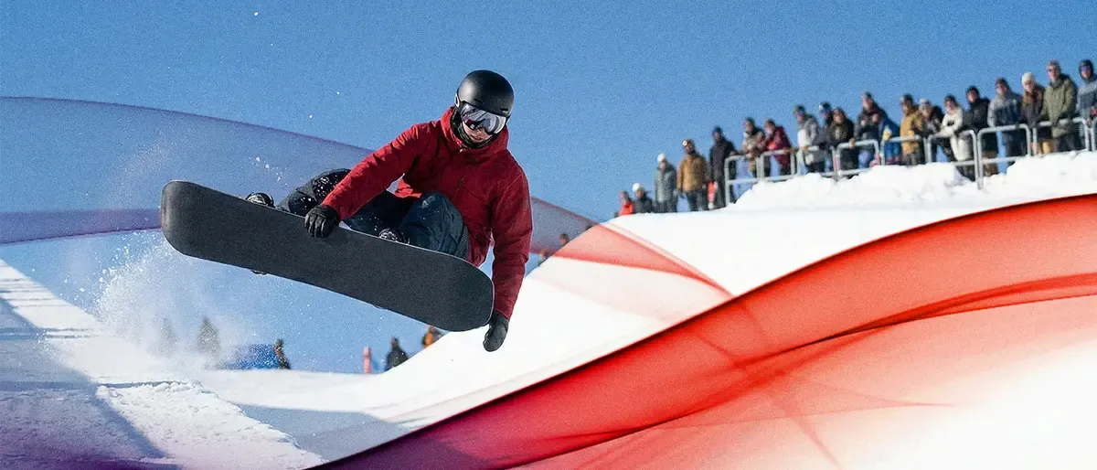 A snowboarder in a red jacket performs an aerial jump out of a snow-covered halfpipe under a clear blue sky while a crowd watches