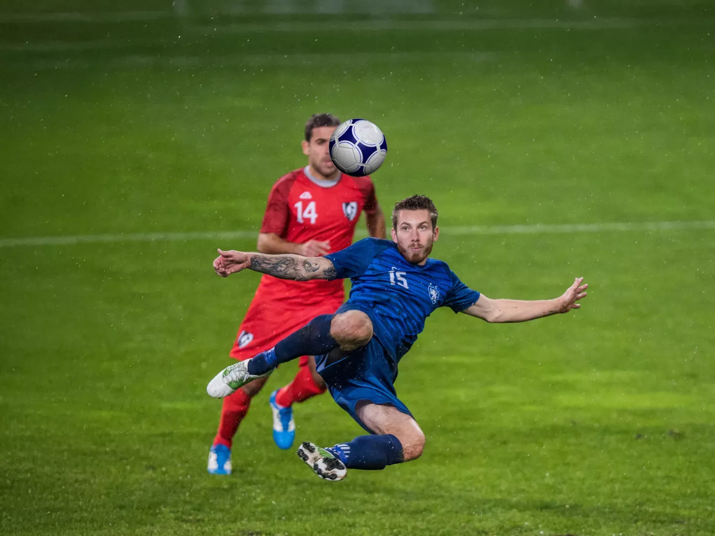 A male soccer player in a blue jersey performs an acrobatic overhead kick on a wet grass field while a defender in a red jersey runs toward him in the background.