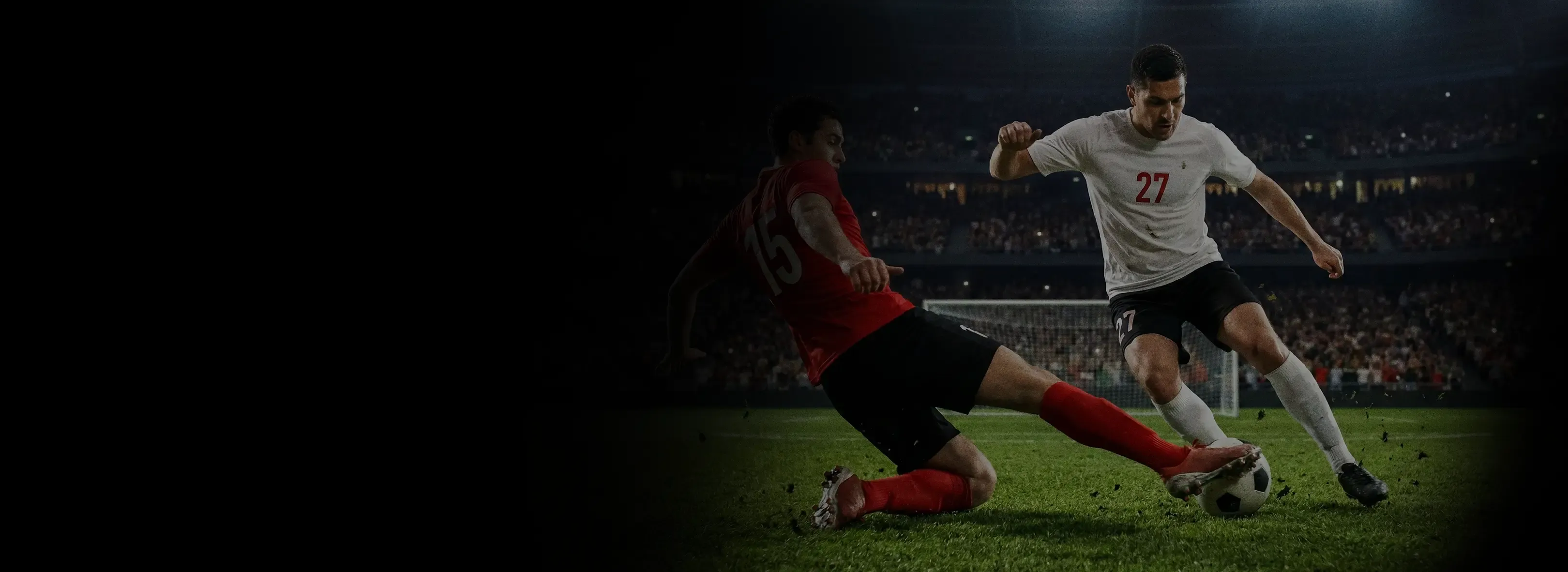 A soccer player in a red jersey performs a slide tackle against an opponent in a white jersey during a stadium match.