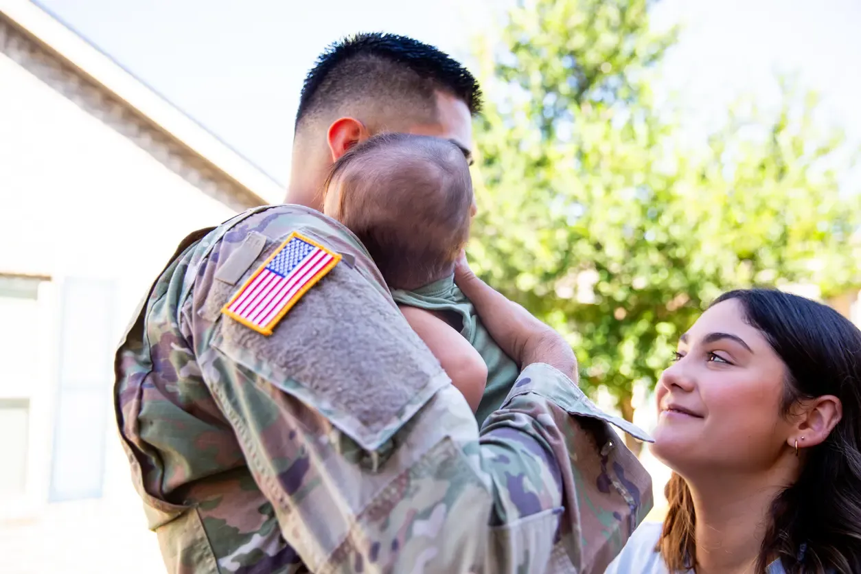 A soldier in camouflage uniform holds his baby while his wife looks on lovingly outdoors.