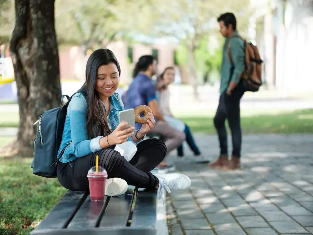 A smiling young woman sits on a park bench using her smartphone while holding a donut.