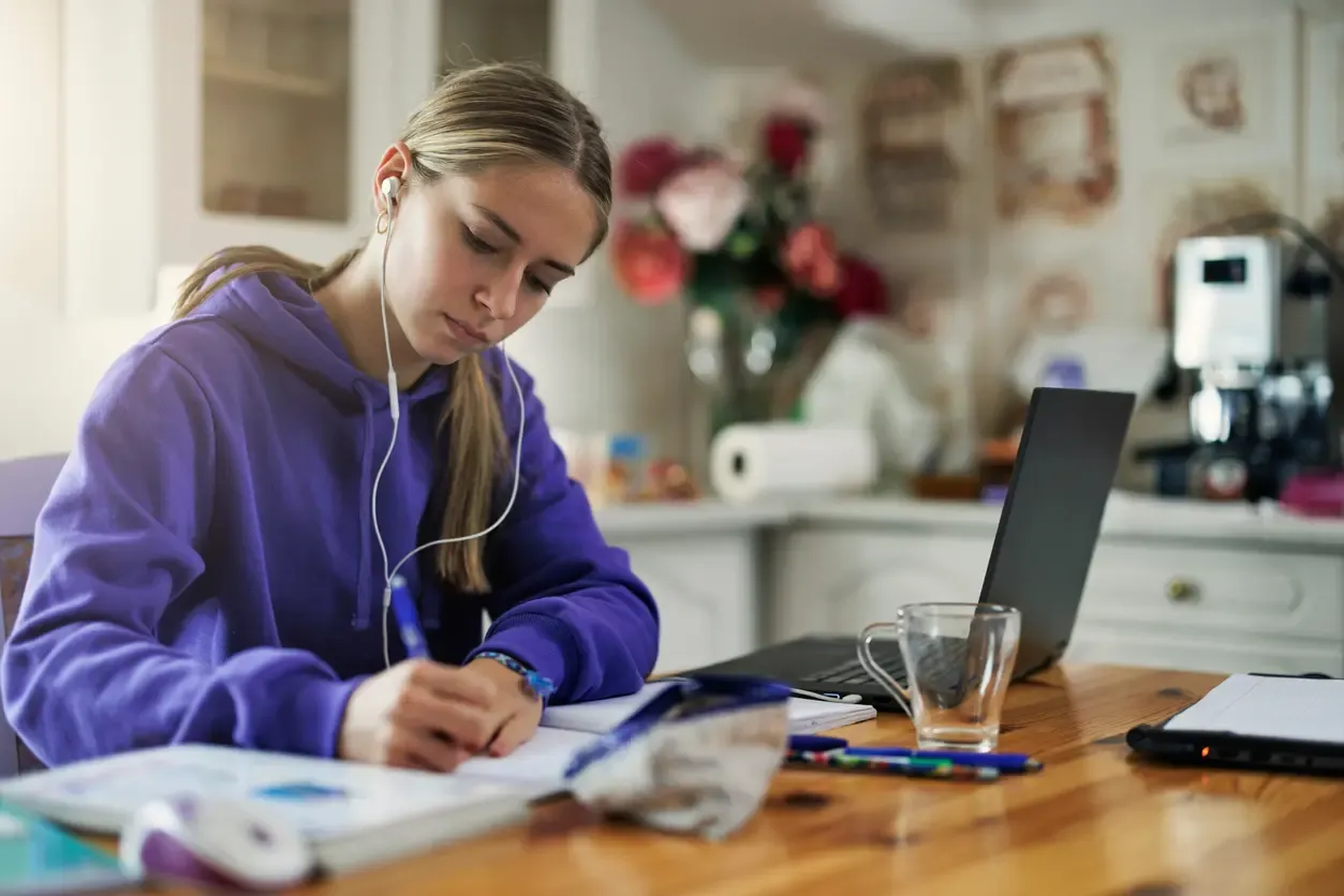 A young woman in a purple hoodie wears earphones while focused on writing in a notebook at a wooden table with a laptop.