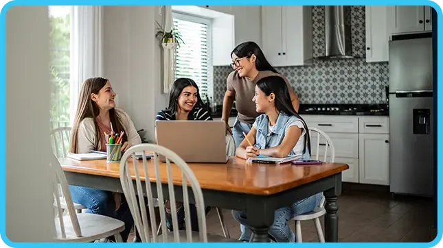Four young women smiling and studying together around a laptop at a kitchen table.
