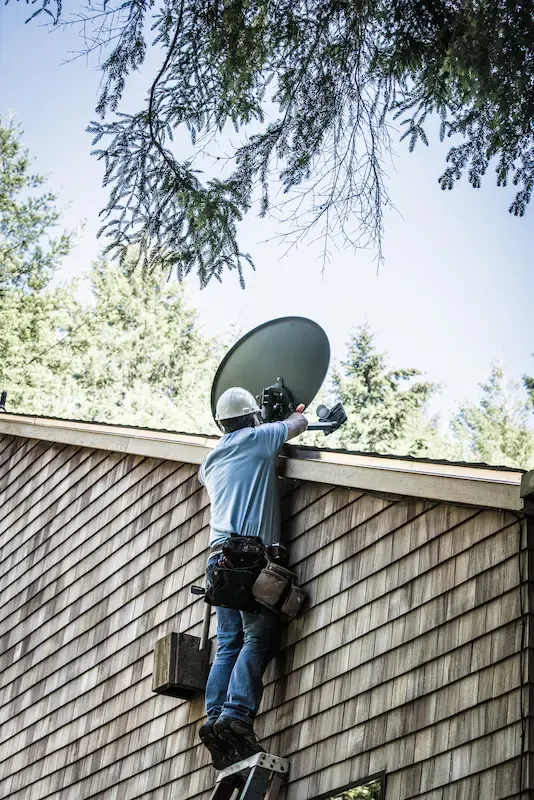 A technician wearing a hard hat and tool belt stands on a ladder to install a satellite dish on the side of a house.