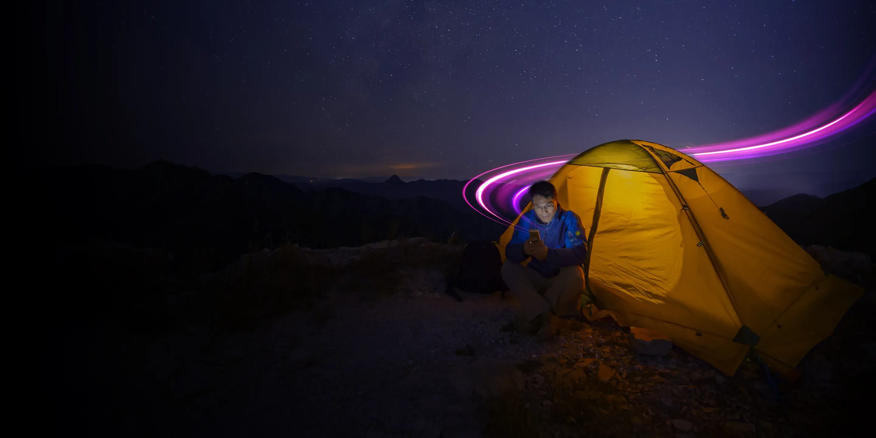 Happer camper illuminated by his cellphone under a stary night sky. He is so happy with his satellite service that he can do anything, anywhere on his phone.