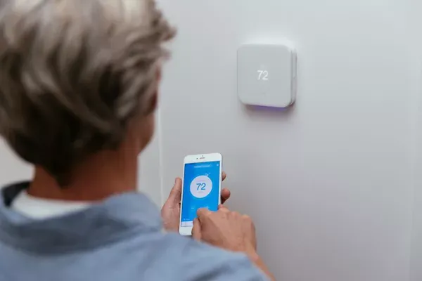 A person stands facing a wall, adjusting a white smart thermostat set to $72^\circ$ on the wall while simultaneously using a mobile app with the same temperature display on their smartphone.