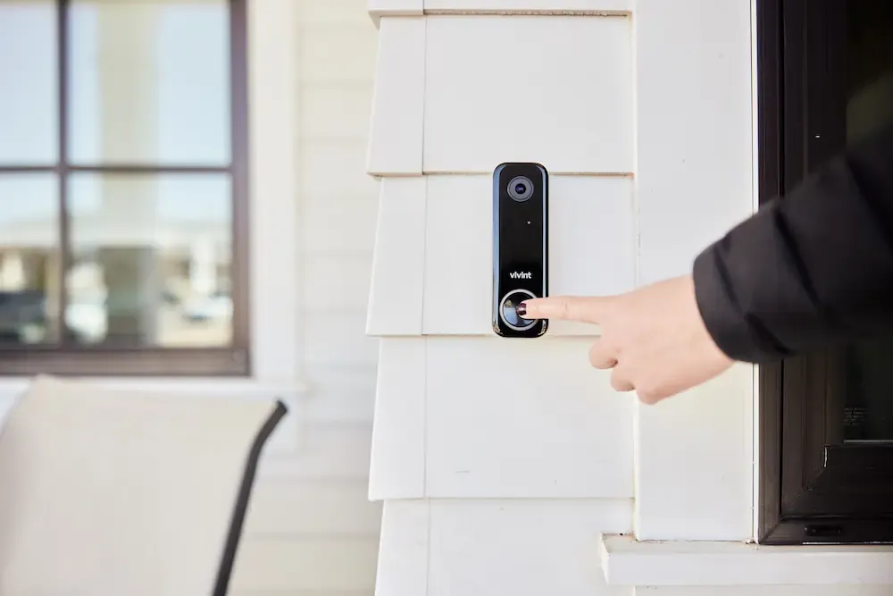 A person presses the button on a Vivint Video Doorbell Camera Pro mounted on the white siding of a home.