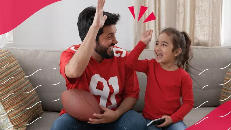 A father and daughter wearing red jerseys give each other a high five while sitting on a couch holding a football, framed by red graphic elements.