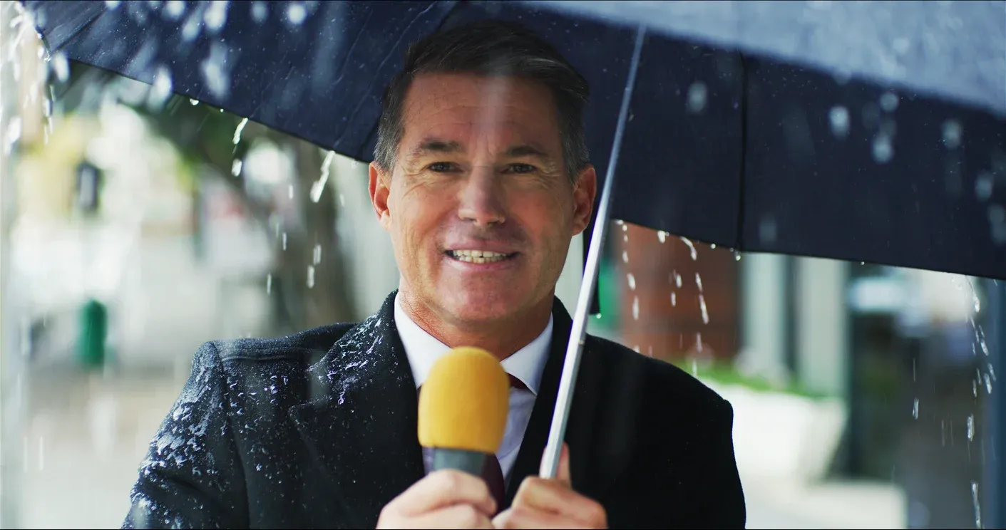 A smiling news reporter holds a microphone and a black umbrella while standing in the rain.