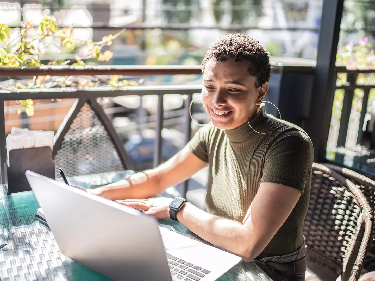 A smiling woman with short hair and large hoop earrings sits at an outdoor table working on a laptop.