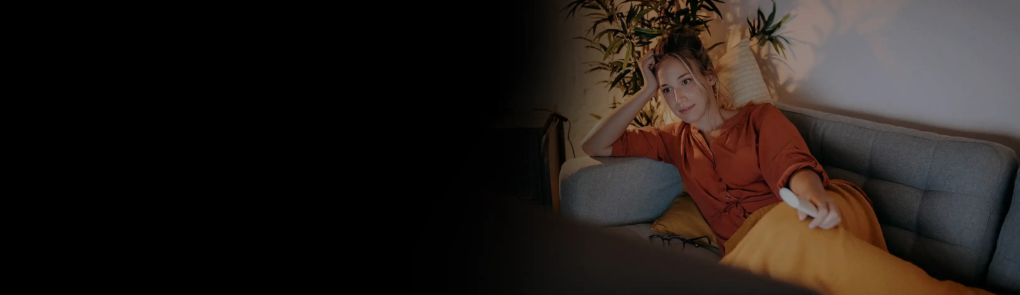 A woman reclines on a gray sofa holding a TV remote in a warmly lit living room at night.